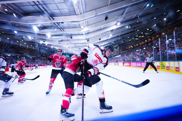 A group of ice hockey players in action on the rink, with one player in white and red being checked by an opponent in red and black The arena is filled with spectators, and the atmosphere is intense under bright lights