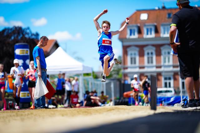 A young athlete in mid-air during a long jump event, surrounded by spectators and officials, with a building and a clear blue sky in the background © Eva Manhart, All rights reserved