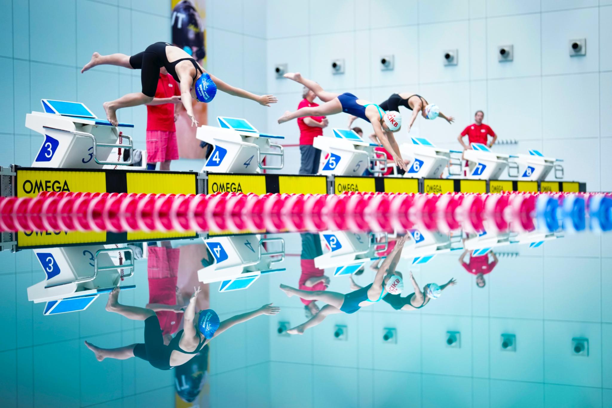 Swimmers diving into a pool from starting blocks, with colorful lane dividers and reflections on the water © Eva Manhart, All rights reserved