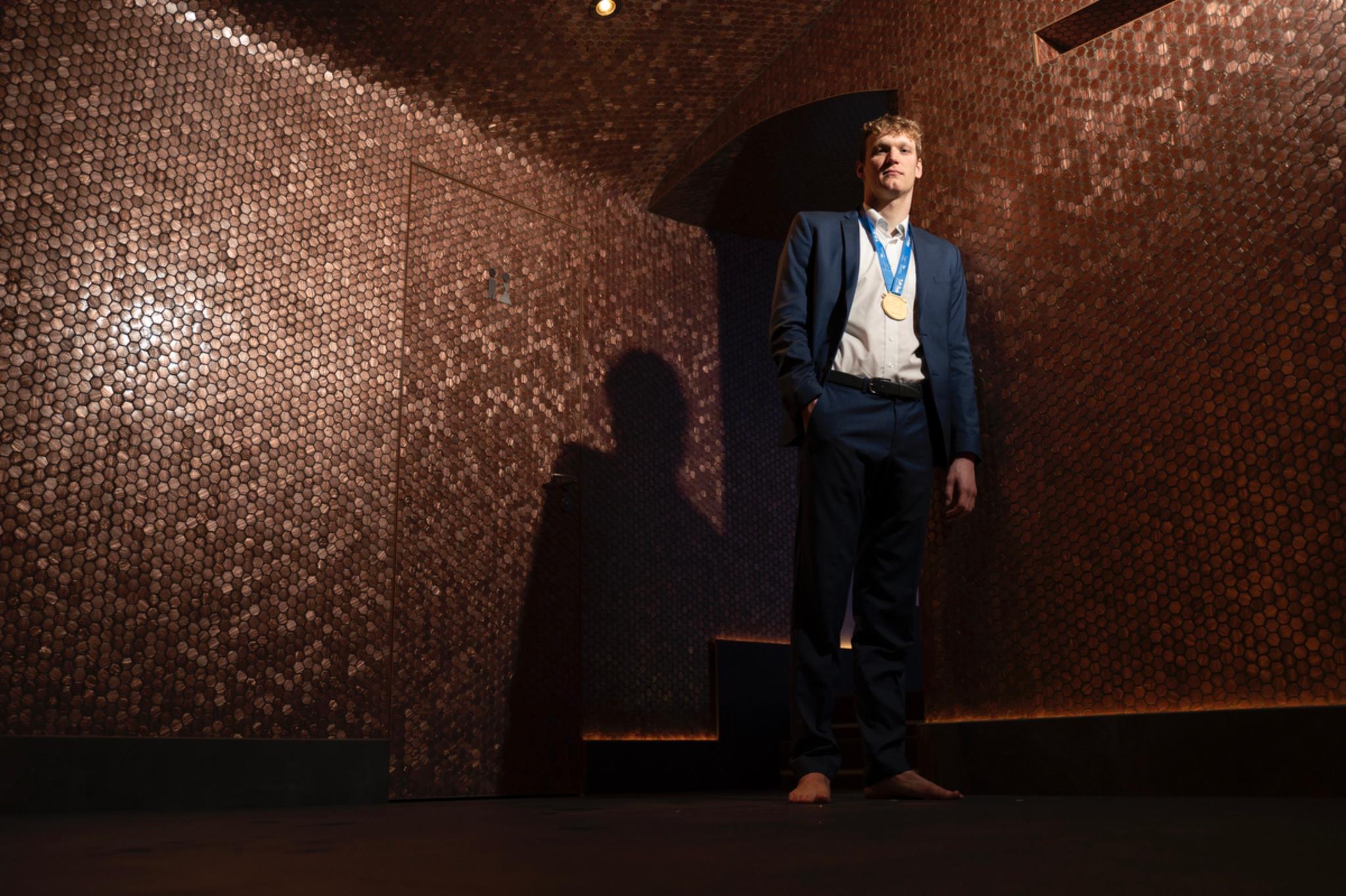 A man in a suit stands barefoot in a dimly lit room with textured walls, casting a shadow behind him