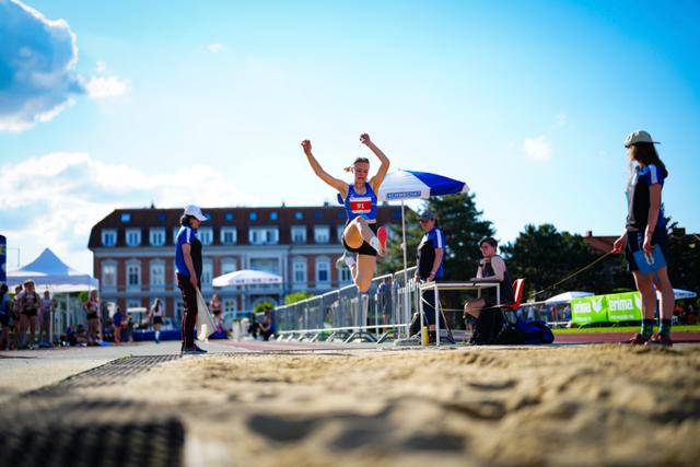 An athlete is mid-air during a long jump event, with officials and spectators in the background on a sunny day © Eva Manhart, All rights reserved