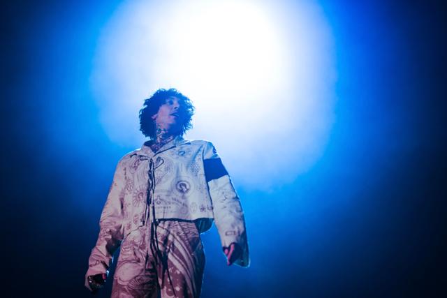 Oliver Sykes with curly hair stands under a bright light, wearing a light-colored jacket with a dark armband, against a blue background