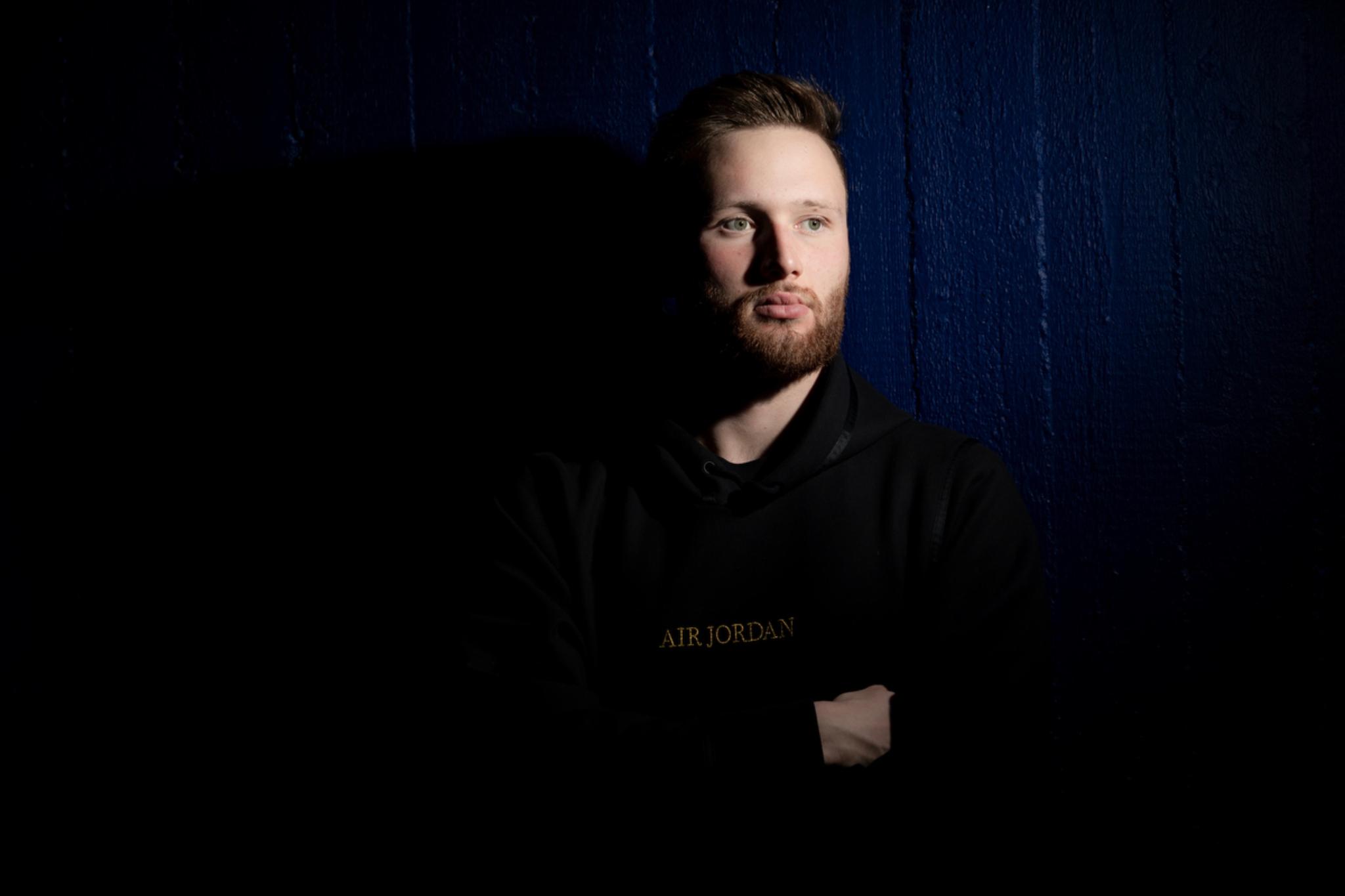 A man with a beard stands against a dark background, partially illuminated by light, wearing a black hoodie