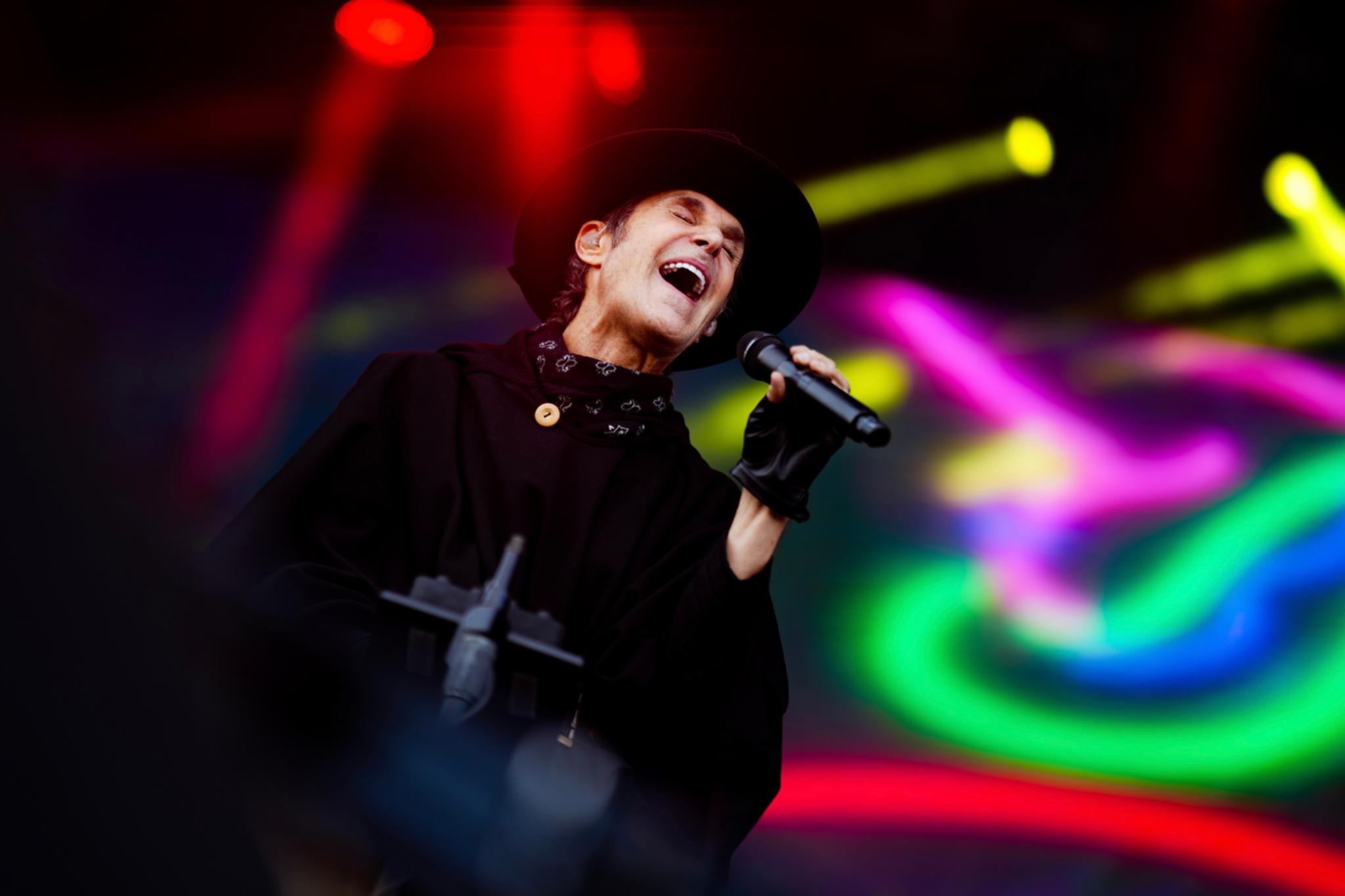 A performer passionately singing into a microphone on stage, wearing a black outfit and hat, with colorful, blurred lights in the background © Eva Manhart, All rights reserved © Eva Manhart, All rights reserved