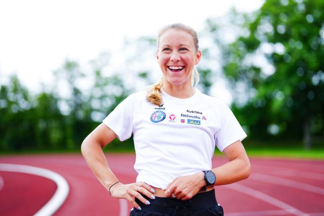 Julia Mayer smiling stands on a running track, wearing a white t-shirt with various logos and black shorts She has a ponytail and is wearing a watch on her left wrist The background features trees and a blurred sky © Eva Manhart, All rights reserved