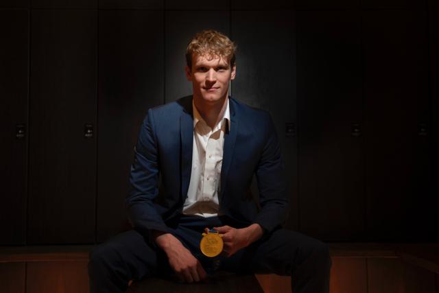 Austrian Swimmer Felix Auböck in a blue suit sits in a dimly lit room, holding a gold medal © Eva Manhart, All rights reserved