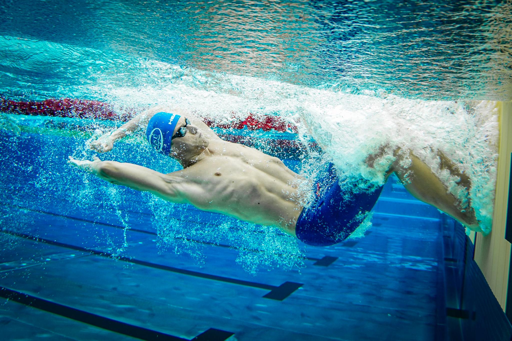 A swimmer in a blue cap and goggles performing a backstroke in a swimming pool, captured underwater © Eva Manhart, All rights reserved