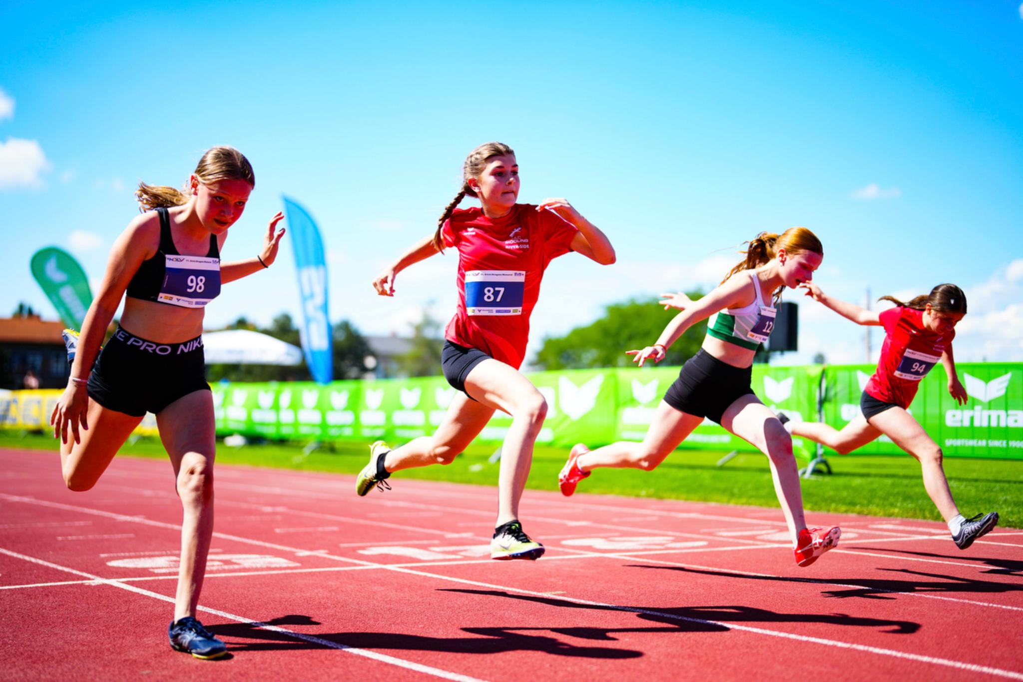 Four young athletes sprinting on a track during a race, with a clear blue sky and green banners in the background © Eva Manhart, All rights reserved