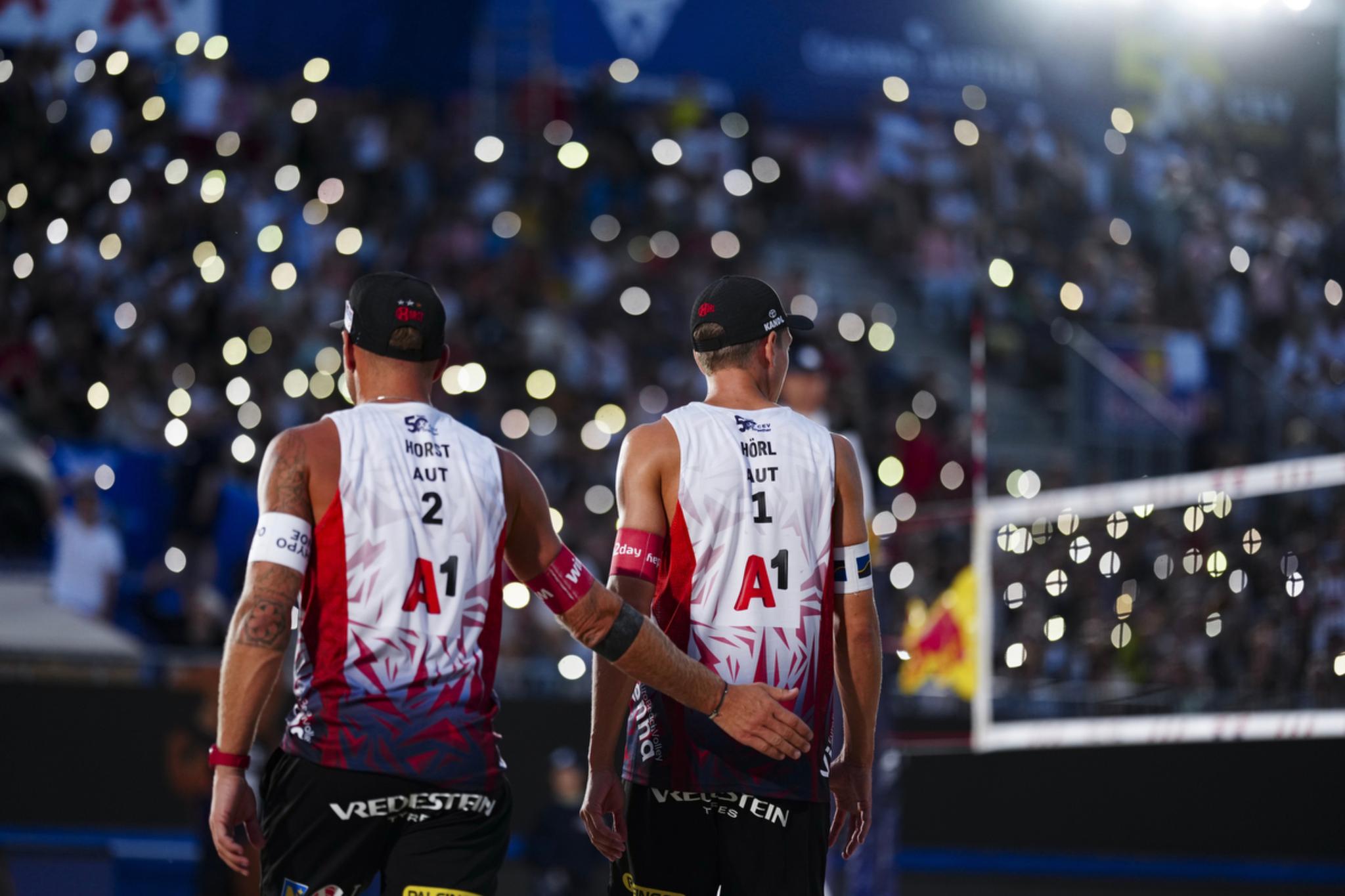 Two beach volleyball players Julian Hörl und Alex Horst in matching uniforms walk side by side on a court, with a crowd of spectators and illuminated lights in the background