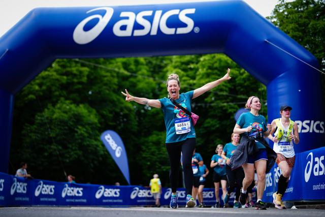 Runners crossing an ASICS-branded finish line, with one person in the foreground celebrating with arms raised © Eva Manhart, All rights reserved