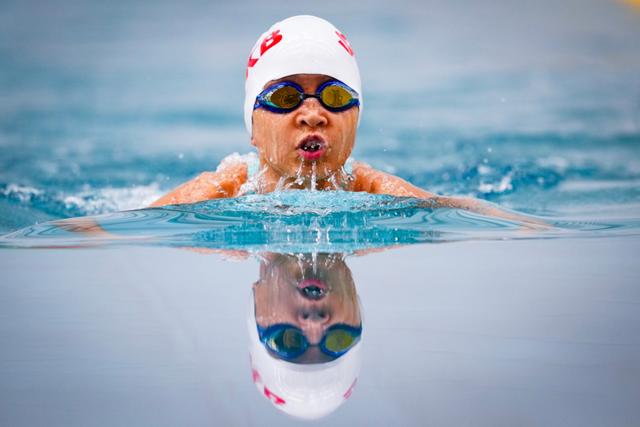 A swimmer wearing a white swim cap and goggles is performing a breaststroke in a pool, with their reflection visible in the water