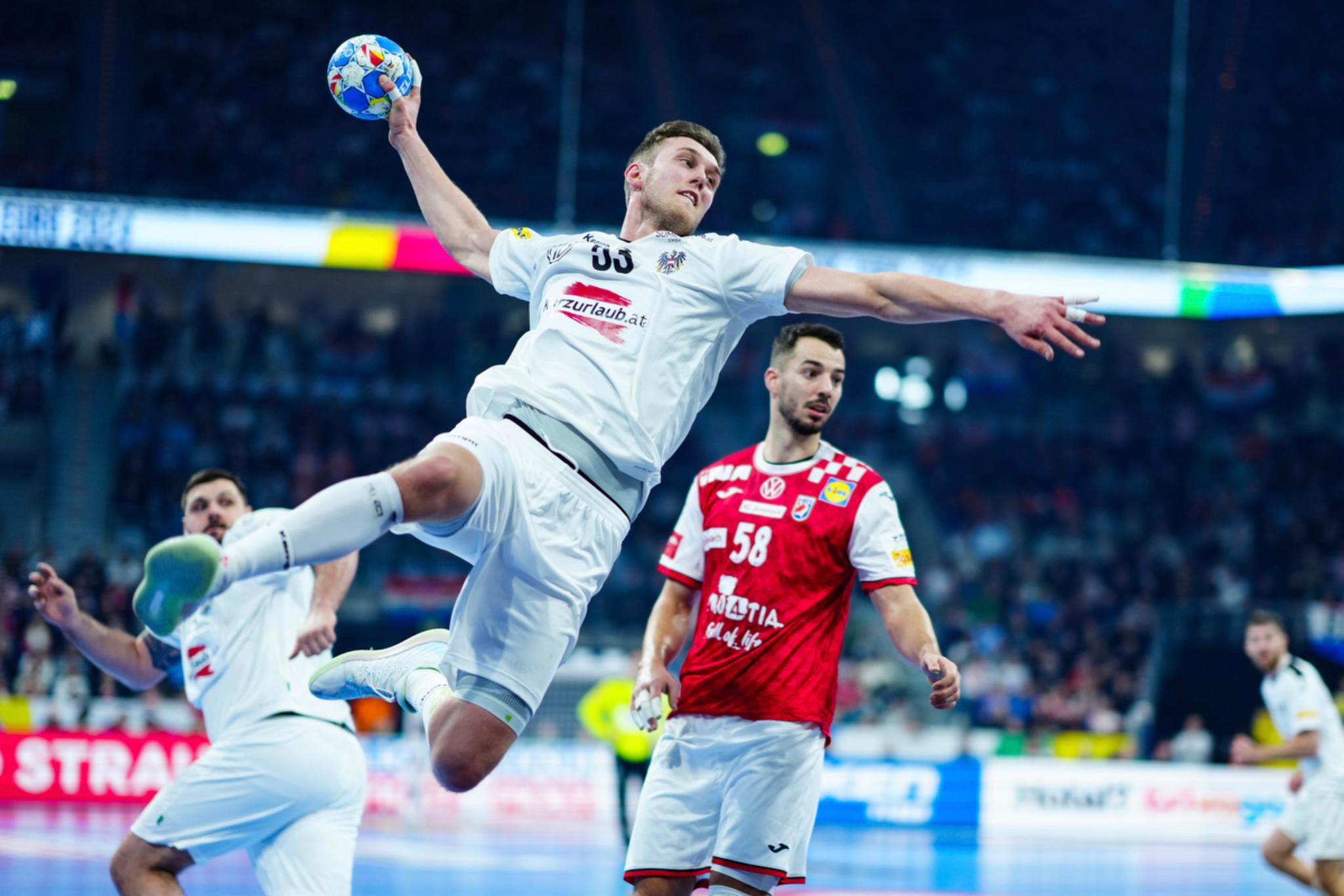 Handball player Mykola Bilyk in a white uniform is leaping into the air to throw the ball, while another player in a red uniform looks on The background features a crowd of spectators and a brightly lit indoor arena © Eva Manhart, All rights reserved