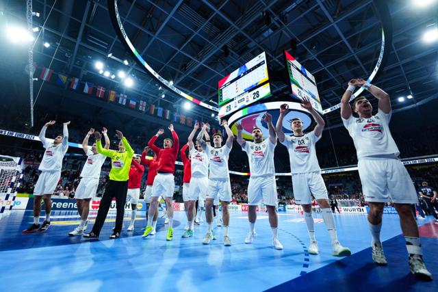 A group of Austrian handball athletes in white uniforms and a few in red celebrate on an indoor sports court, raising their arms in victory The background features a large scoreboard and a crowd of spectators © Eva Manhart, All rights reserved