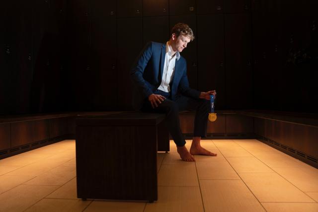 Austrian swimmer Felix Auböck in a suit sits barefoot in a dimly lit room, holding a gold medal © Eva Manhart, All rights reserved