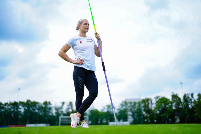 Victoria Hudson standing on a grassy field, holding a javelin upright, with a cloudy sky and trees in the background © Eva Manhart, All rights reserved