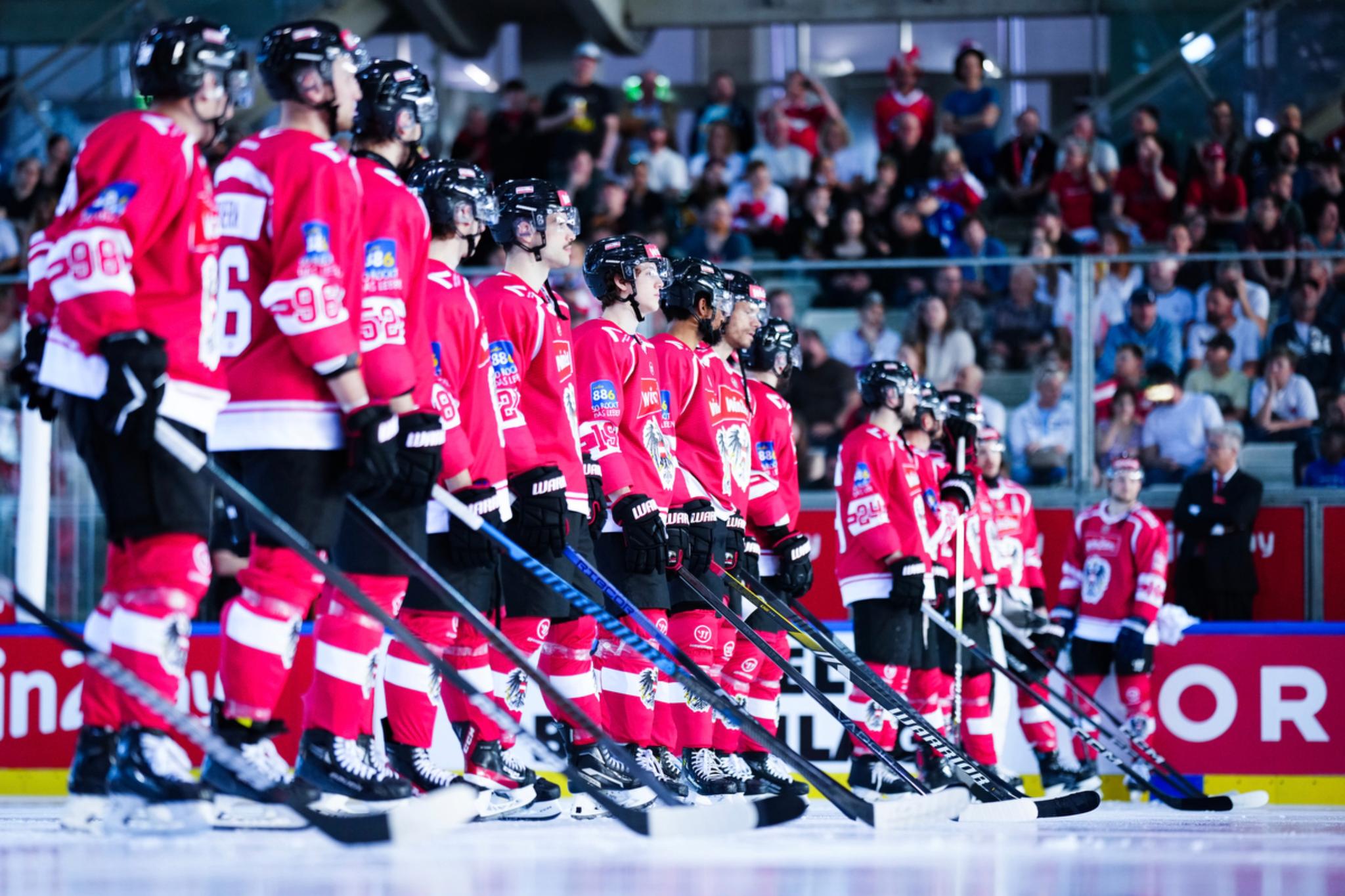 A hockey team in red jerseys stands in a line on the ice, holding their sticks, with a crowd of spectators in the background