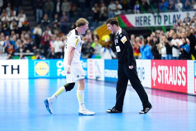 A handball player in a white uniform and a coach in a black outfit are walking on a court, with a crowd of spectators in the background