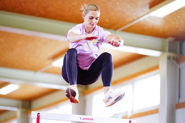 Victoria Hudson in athletic attire is mid-jump over a hurdle in an indoor setting