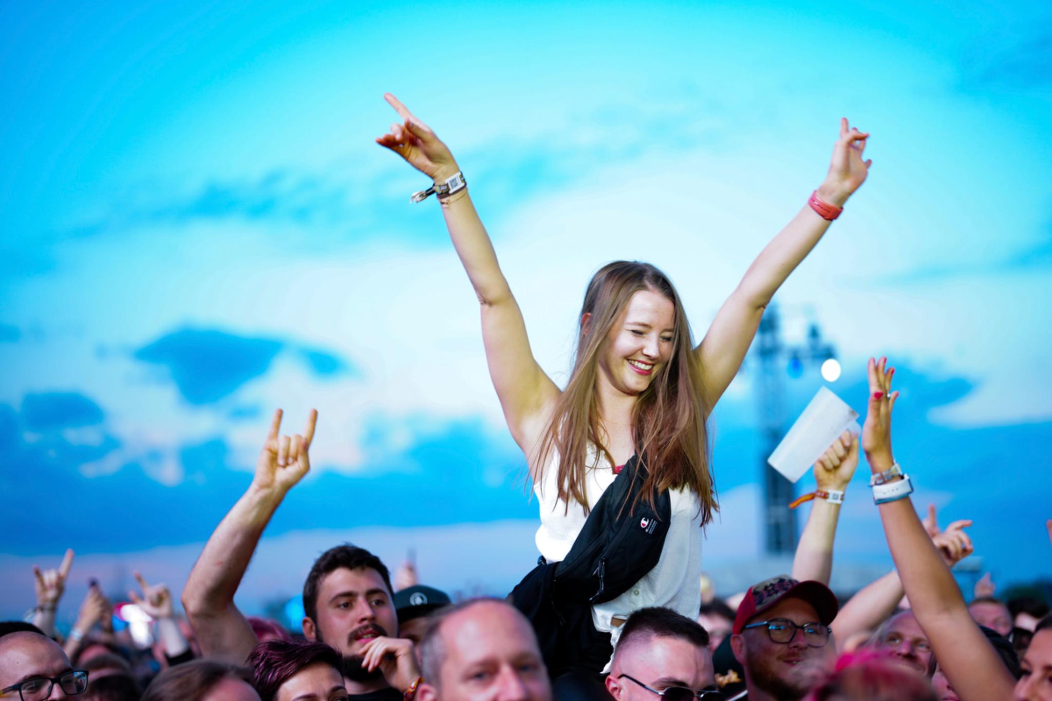 A young woman is being lifted above a crowd at an outdoor event, smiling and raising her arms in excitement The sky is blue with scattered clouds in the background