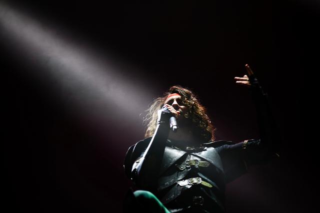 A performer with long hair sings passionately into a microphone under a dramatic spotlight on a dark stage © Eva Manhart, All rights reserved