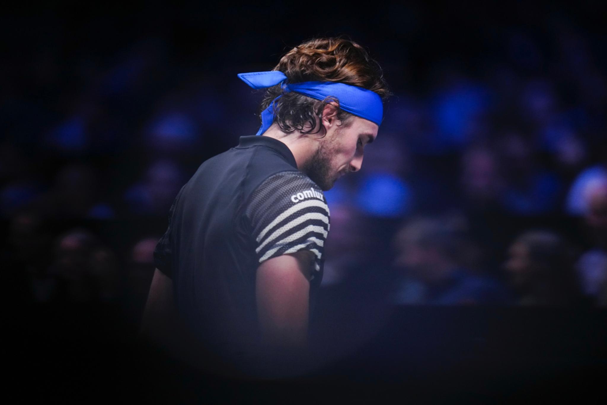 Tennis player Stefanos TsitsIpas wearing a blue headband and a dark outfit, standing in a dimly lit environment with a blurred audience in the background © Eva Manhart, All rights reserved