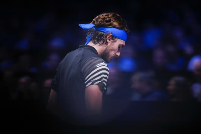 Tennis player Stefanos TsitsIpas wearing a blue headband and a dark outfit, standing in a dimly lit environment with a blurred audience in the background © Eva Manhart, All rights reserved