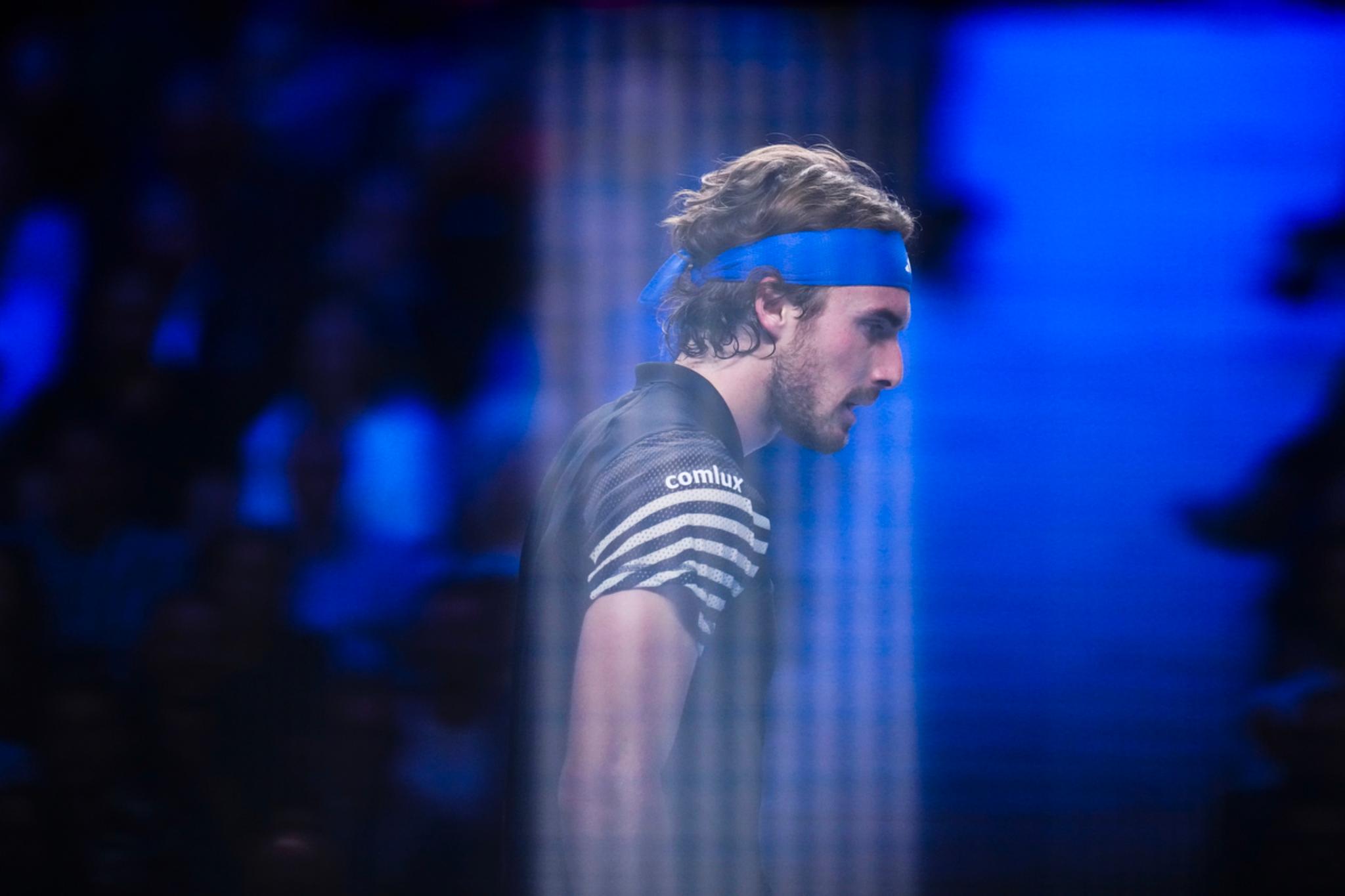 Tennis Player Stefanos Tsitsipas wearing a blue headband and a striped shirt, captured in a side profile with a blurred, blue-toned background