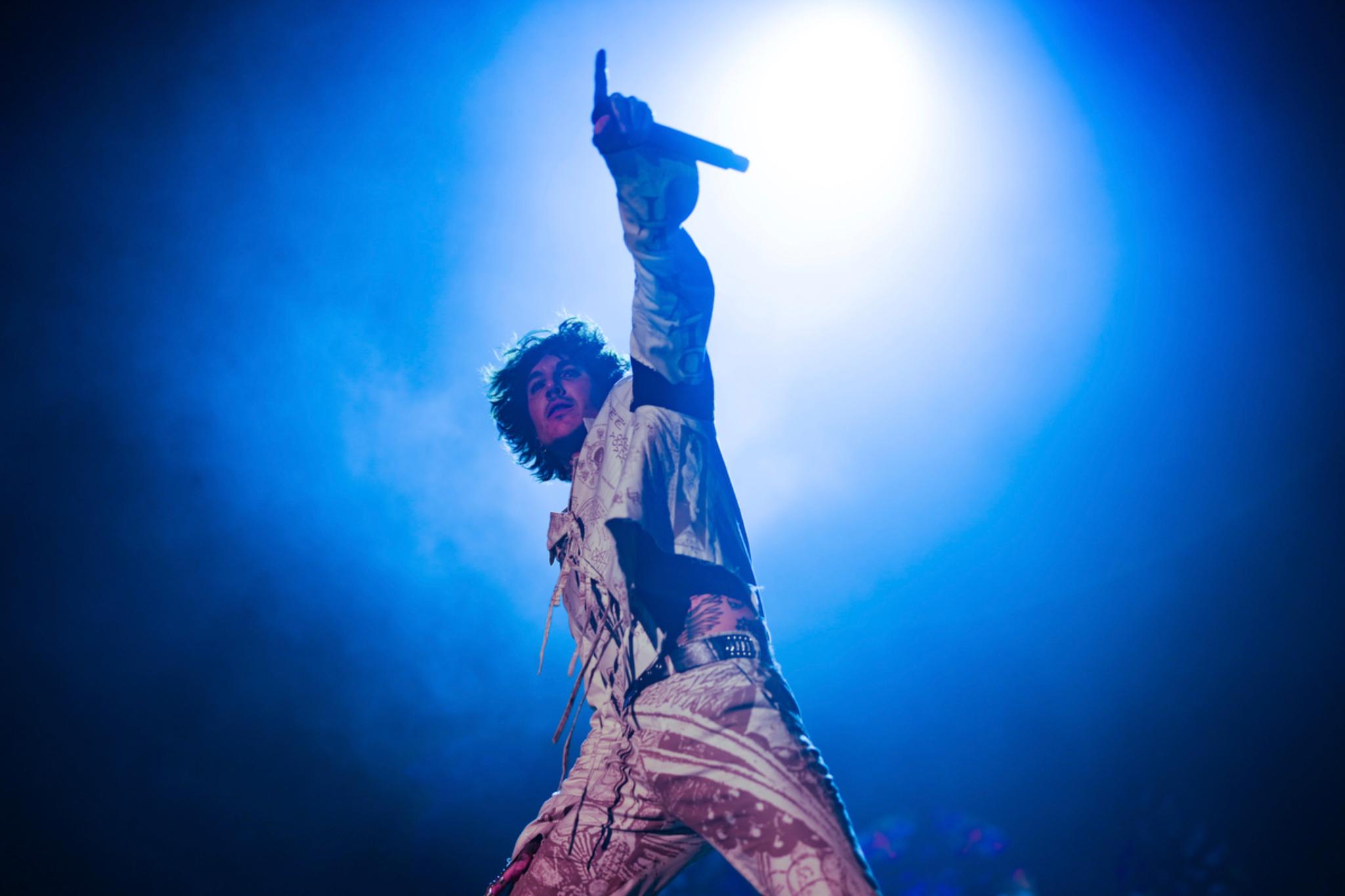 Oliver Sykes on stage, illuminated by a bright blue spotlight, raises one arm while holding a microphone