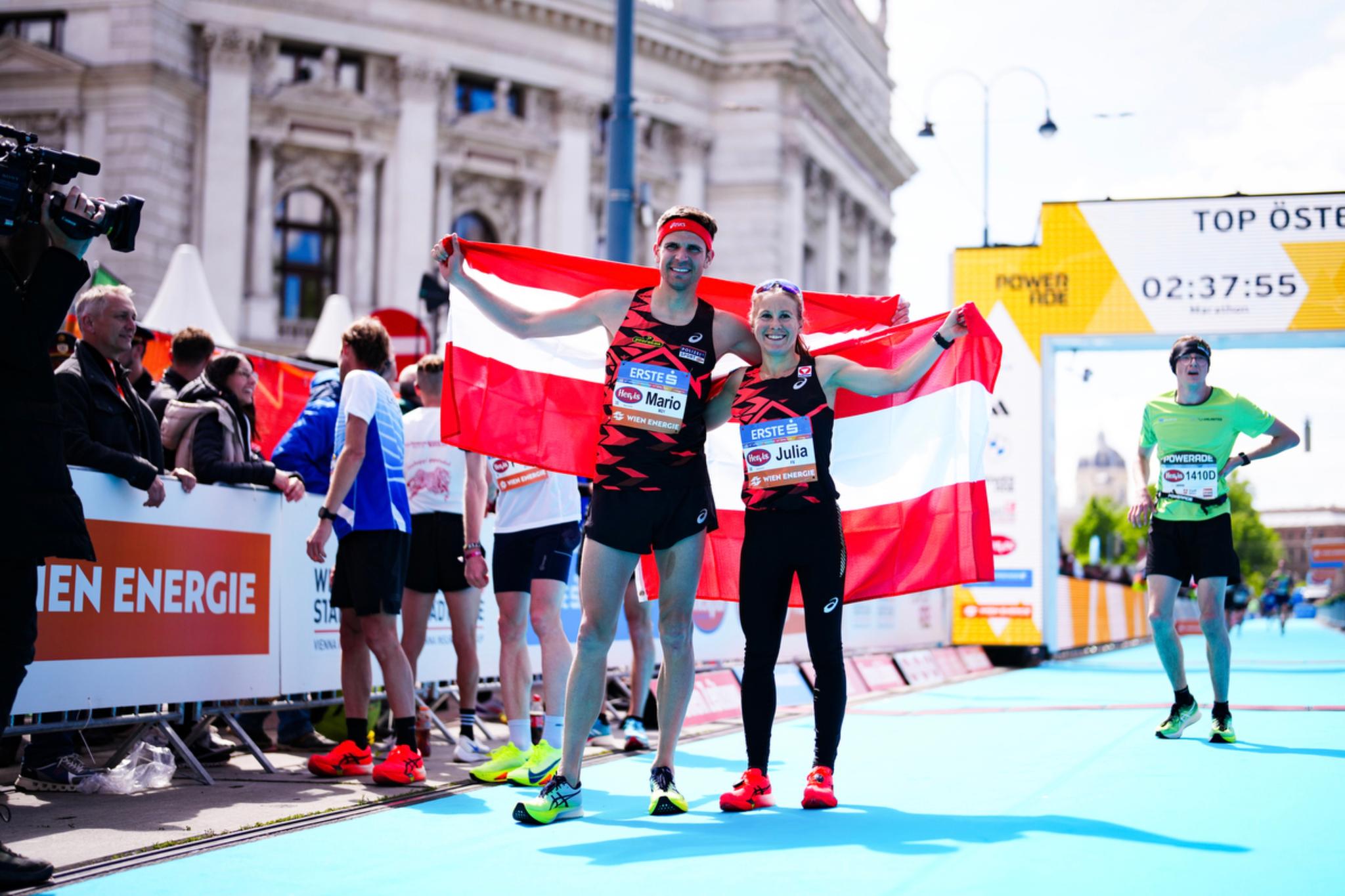 Two athletes holding an Austrian flag celebrate at a marathon finish line, surrounded by other runners and spectators