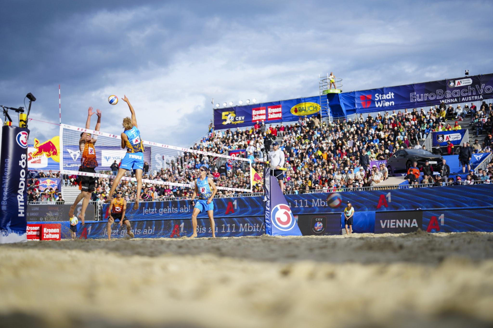 A beach volleyball match in progress with players jumping near the net, a large crowd of spectators in the stands, and a cloudy sky overhead