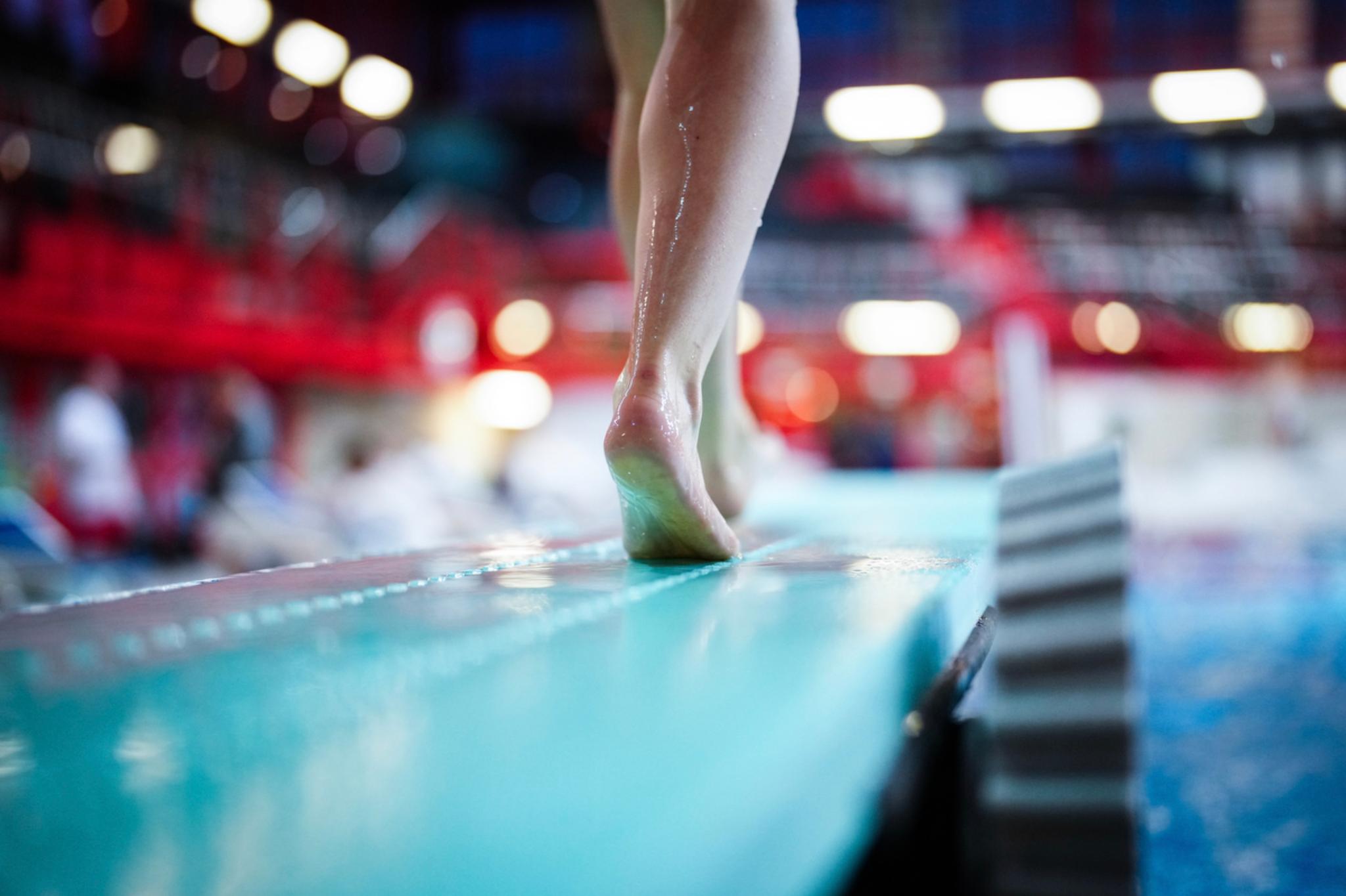 A person standing on a diving board, with a blurred background of an indoor swimming pool area