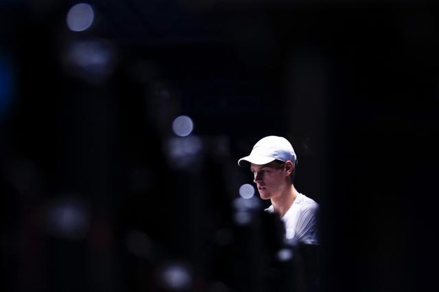 Tennis Player Jannik Sinner wearing a white cap, partially illuminated in a dark setting with blurred foreground elements © Eva Manhart, All rights reserved