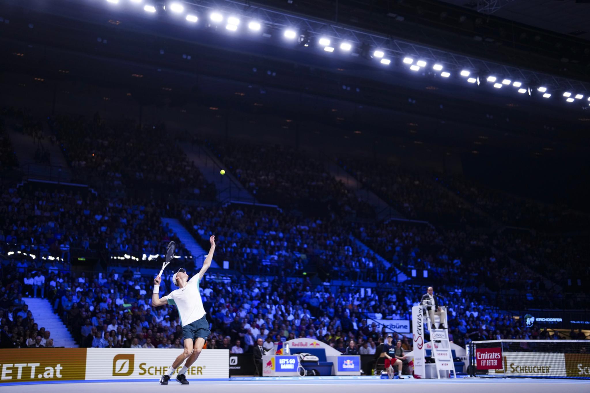 Ttennis player Jannik Sinner is serving on an indoor court with a large audience in the background and bright overhead lights illuminating the scene