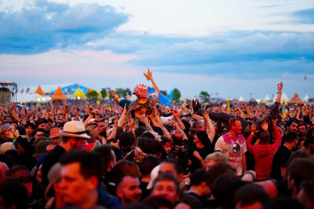 A large crowd of people at an outdoor event, with some individuals raising their hands and one person being lifted above the crowd The background features a cloudy sky and colorful tents