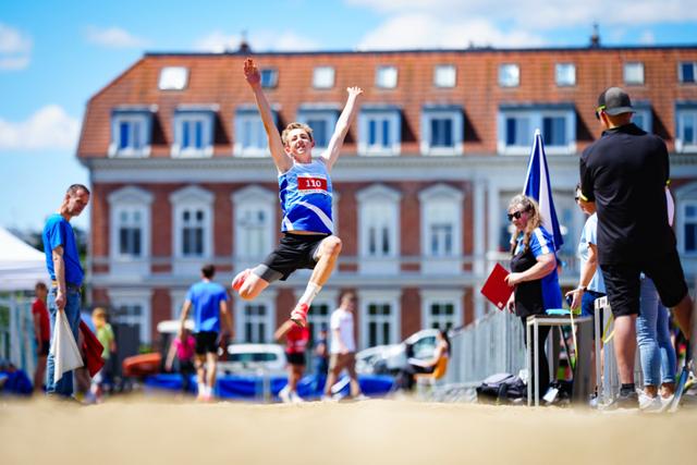 A young athlete in mid-air during a long jump event, surrounded by spectators and officials, with a building in the background © Eva Manhart, All rights reserved