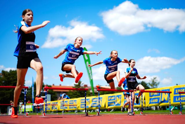 Four young athletes in blue uniforms are mid-air while jumping over hurdles on a track under a bright blue sky with scattered clouds © Eva Manhart, All rights reserved