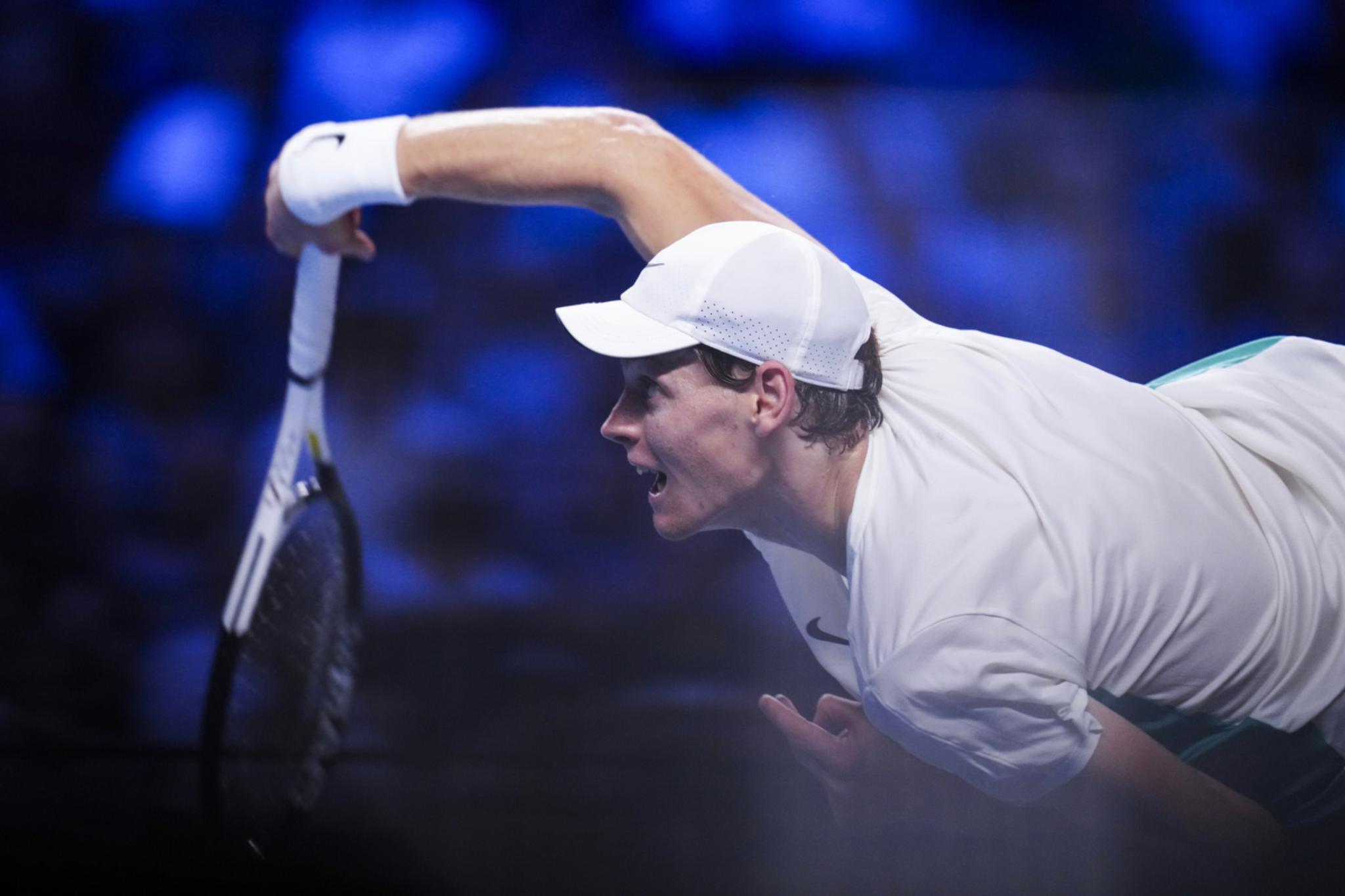 Tennis player Jannik Sinner in a white outfit and cap, reaching out with a racket to hit a ball, with a blurred blue background