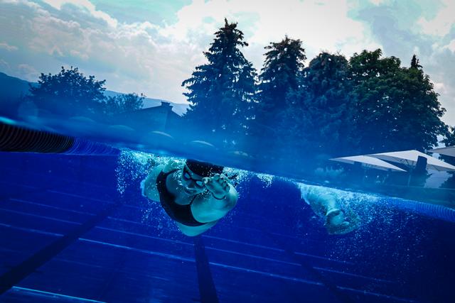 A swimmer underwater in a pool, with trees and a cloudy sky visible above the water's surface