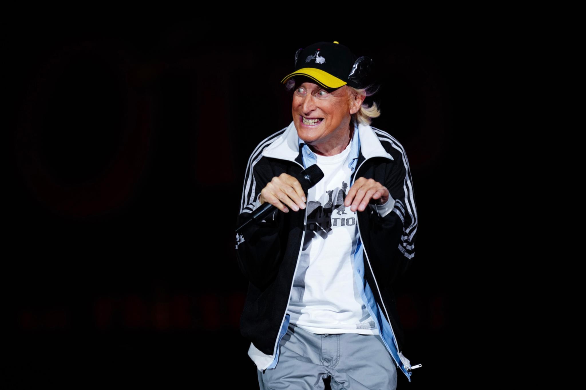 Otto Waalkes wearing a black and white jacket, a white shirt, and a yellow and black cap, standing against a dark background and making a playful gesture with their hands © Eva Manhart, All rights reserved