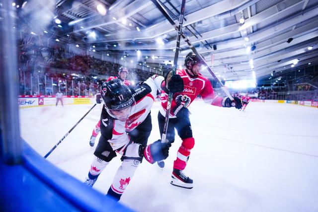 Two ice hockey players in action, one in a red jersey and the other in a white jersey, engaged in a physical battle near the rink's glass barrier, with a blurred crowd and bright arena lights in the background